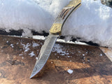 Handmade Damascus Steel Stag Engraved Bolster Folding knife with a gold handle on a snowy surface