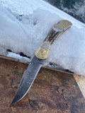 Handmade Damascus Steel Stag Engraved Bolster Folding knife with a gold and stag handle on a snowy surface