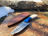 Damascus steel hunting & skinning knife with a camel bone handle and leather sheath on a brown tile with a wooden log and snow in the background